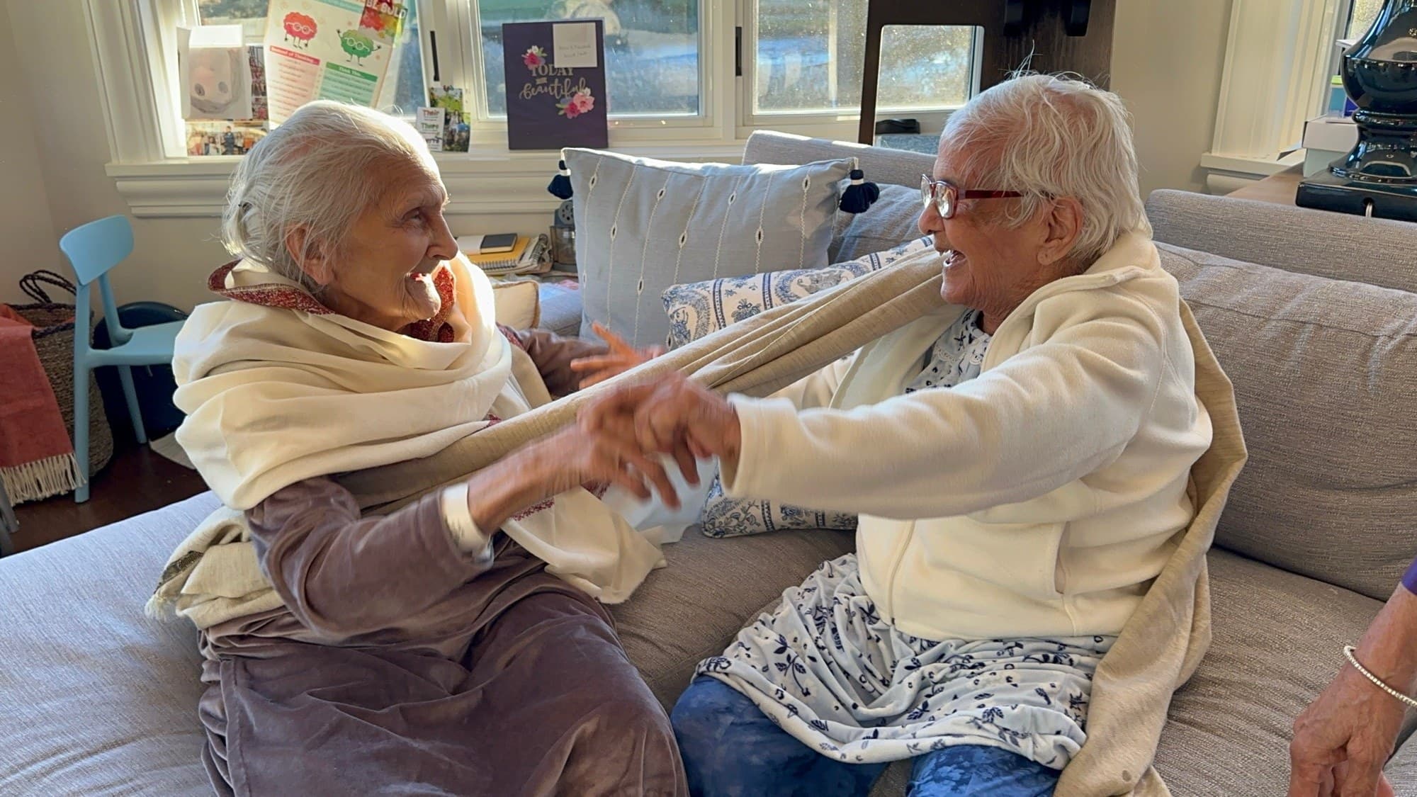 Two older women sitting together on a sofa, holding hands and laughing
