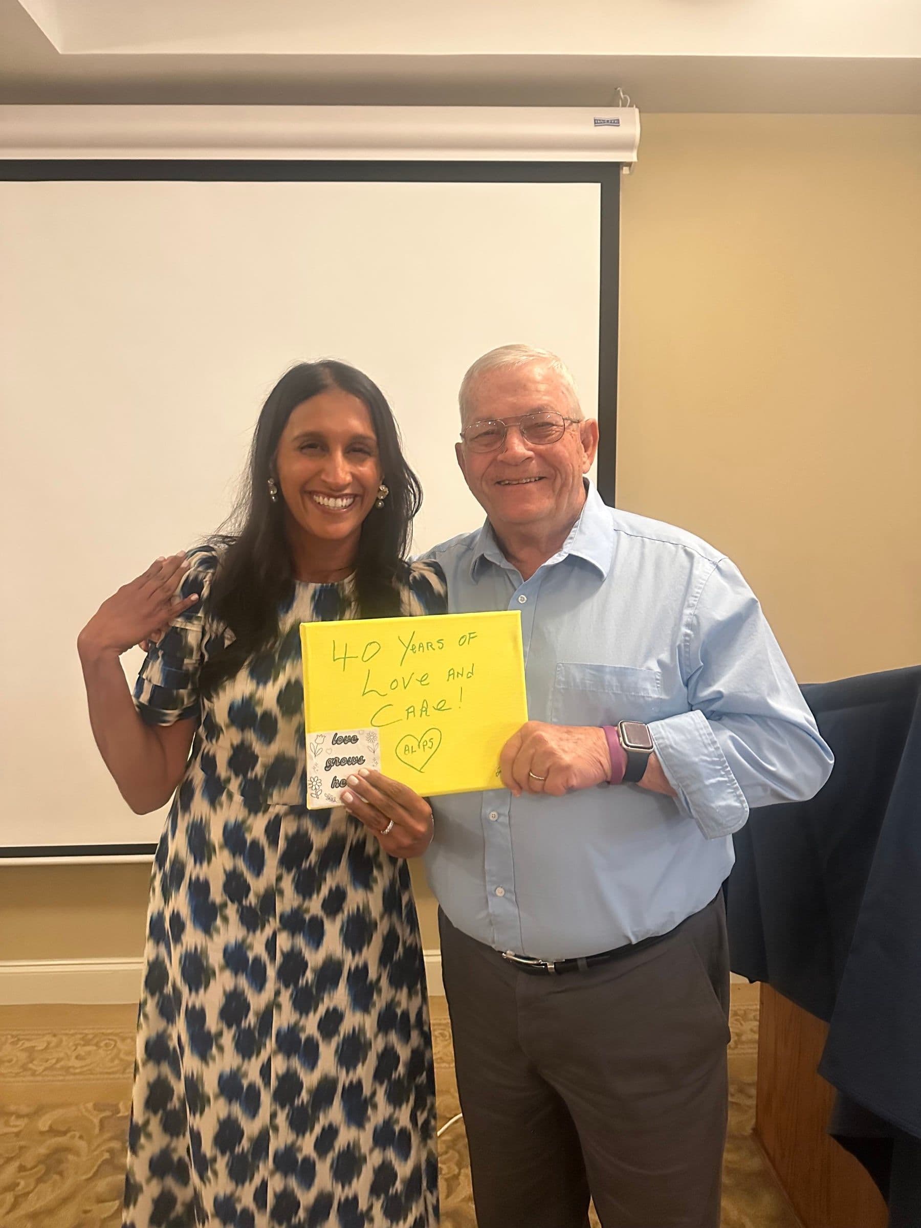 Dr. Tina Sadarangani standing arm in arm with an older man at a conference; together they hold up a handmade yellow sign reading “40 Years of Love and Care!”