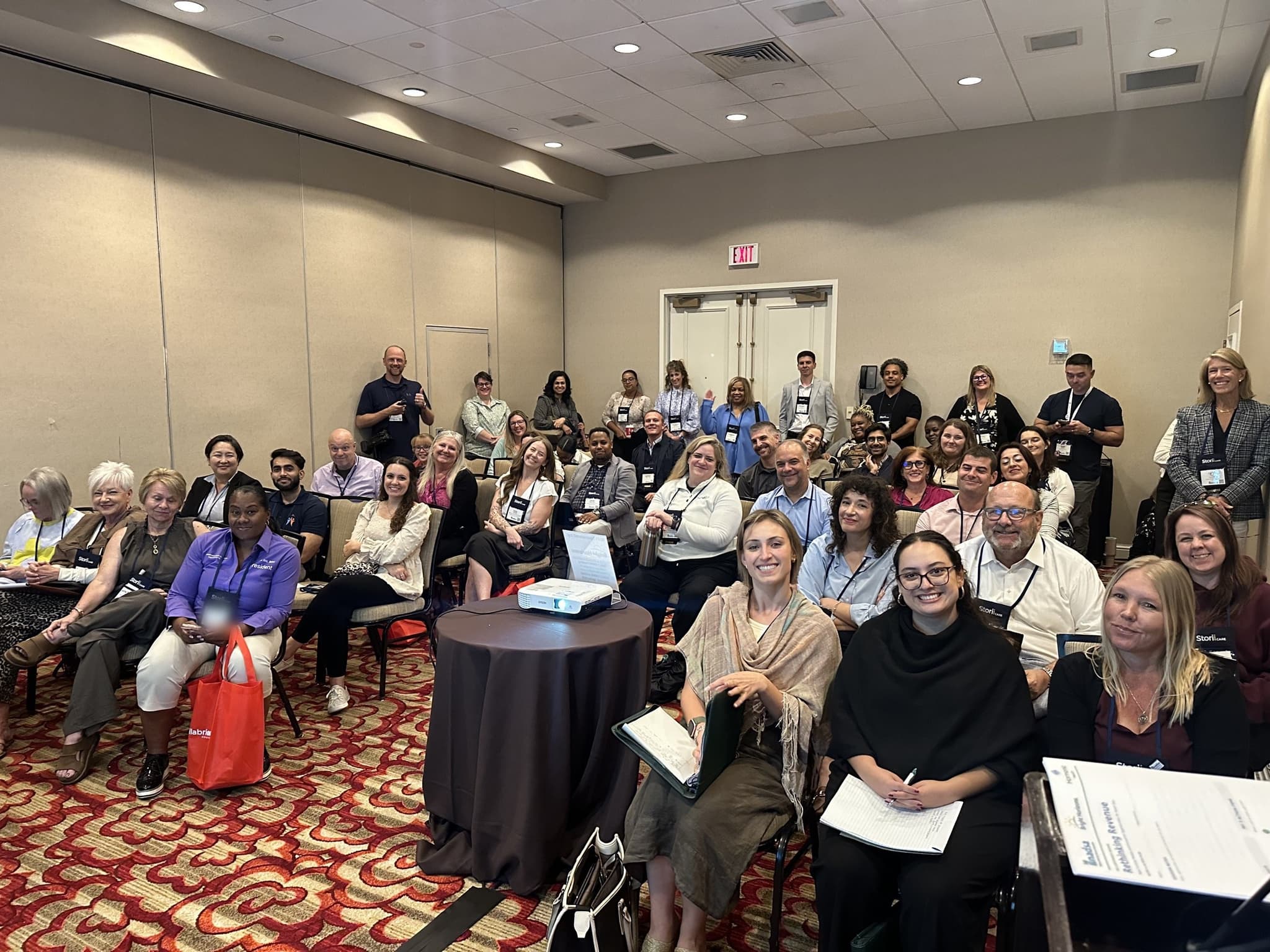 About forty people seated and standing together in a conference room, turned toward the camera and smiling, at a session on adult day services
